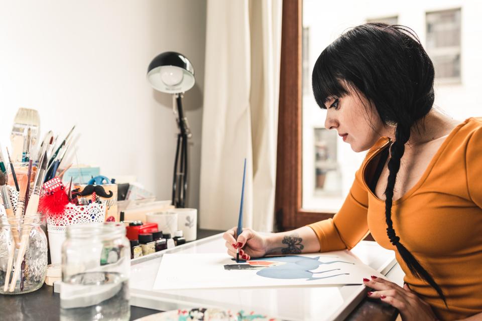 Illustrator Fatinha Ramos zit aan de tekentafel. Voor haar ligt een blad papier met een half afgewerkte illustratie, die ze met haar rechterhand verder afwerkt.